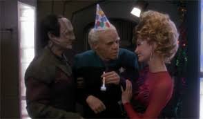Garak and a woman holding up an age Bashir in a birthday hat